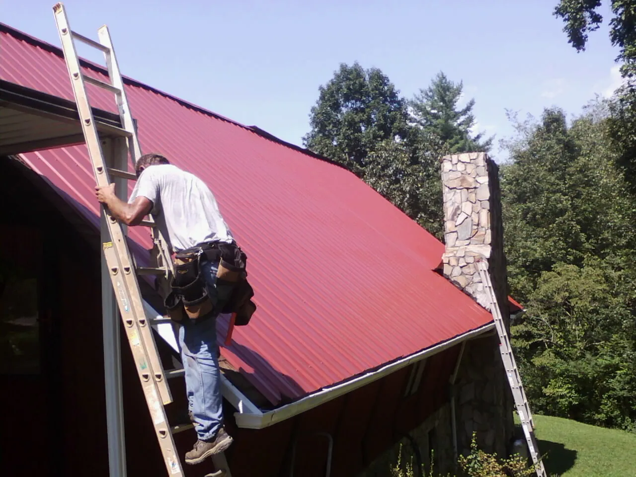 Roofer on a ladder beside a red metal roof and stone chimney — Soffit & Fascia Repair by Roofing Brothers in Botkins